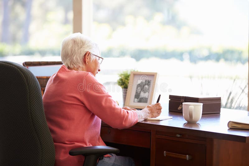 Senior Woman Writing Memoirs In Book At Desk With Hot Drink