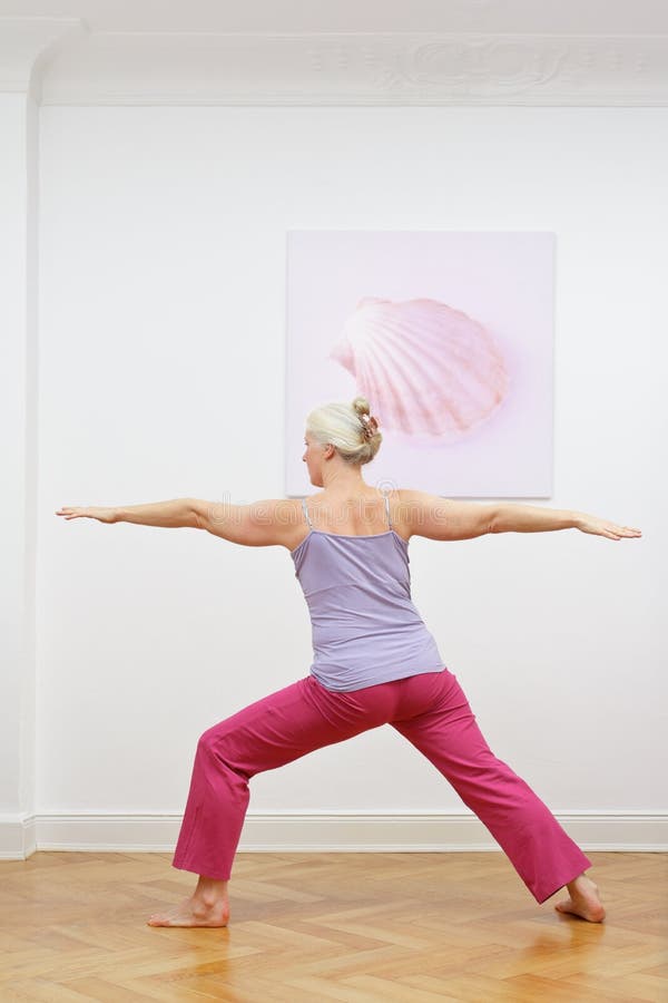 Senior woman with gray hair doing yoga exercises at home in front of a wall with a picture, asana warrior II. Senior woman with gray hair doing yoga exercises at home in front of a wall with a picture, asana warrior II