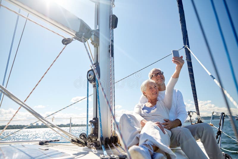 Sailing, technology, tourism, travel and people concept - happy senior couple with smartphone taking selfie on sail boat or yacht deck floating in sea. Sailing, technology, tourism, travel and people concept - happy senior couple with smartphone taking selfie on sail boat or yacht deck floating in sea