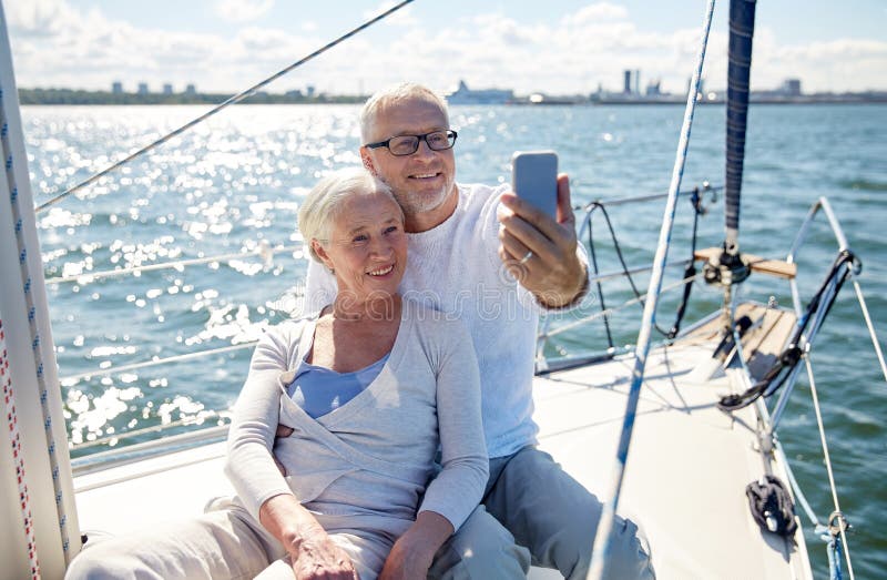 Sailing, technology, tourism, travel and people concept - happy senior couple with smartphone taking selfie on sail boat or yacht deck floating in sea. Sailing, technology, tourism, travel and people concept - happy senior couple with smartphone taking selfie on sail boat or yacht deck floating in sea