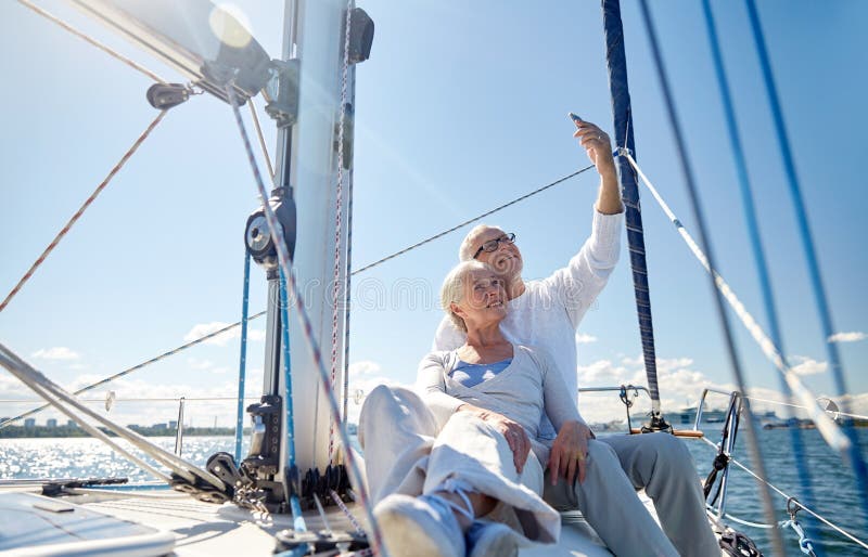 Sailing, technology, tourism, travel and people concept - happy senior couple with smartphone taking selfie on sail boat or yacht deck floating in sea. Sailing, technology, tourism, travel and people concept - happy senior couple with smartphone taking selfie on sail boat or yacht deck floating in sea