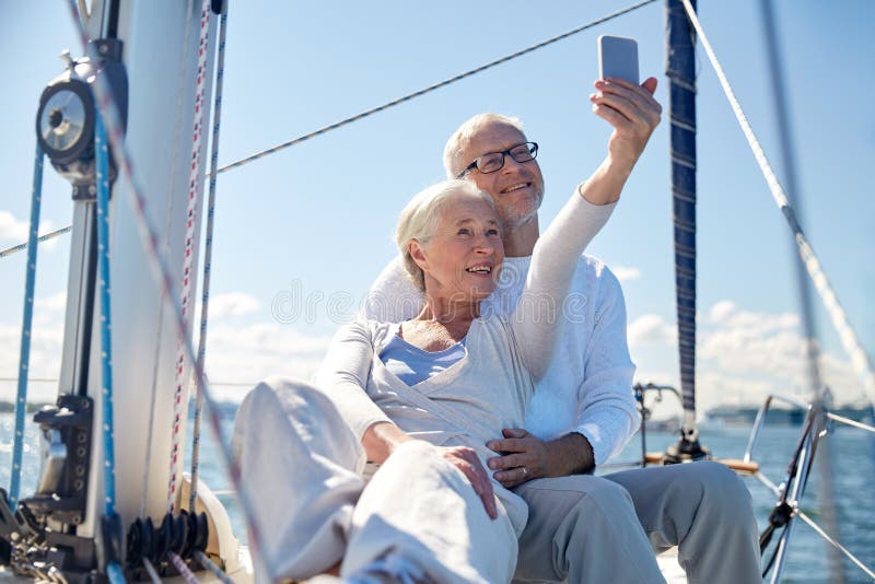 Sailing, technology, tourism, travel and people concept - happy senior couple with smartphone taking selfie on sail boat or yacht deck floating in sea. Sailing, technology, tourism, travel and people concept - happy senior couple with smartphone taking selfie on sail boat or yacht deck floating in sea