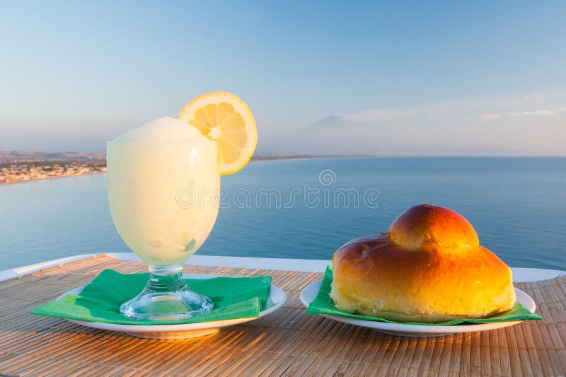 Sicilian lemon granita and a typical warm brioche with blue sea and Mount etna in the background. Sicilian lemon granita and a typical warm brioche with blue sea and Mount etna in the background