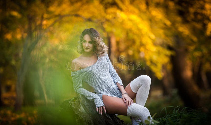 Sensual girl with long legs sitting on a stump in an autumnal scene.Long legs attractive blonde with curly hair relaxing