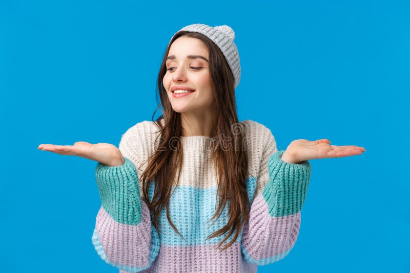 Sensual, cute and tender feminine young woman in sweater, winter hat, raising hands, holding products on palms, smiling making choice, have two good gifts, special discount offers, blue background. Sensual, cute and tender feminine young woman in sweater, winter hat, raising hands, holding products on palms, smiling making choice, have two good gifts, special discount offers, blue background.