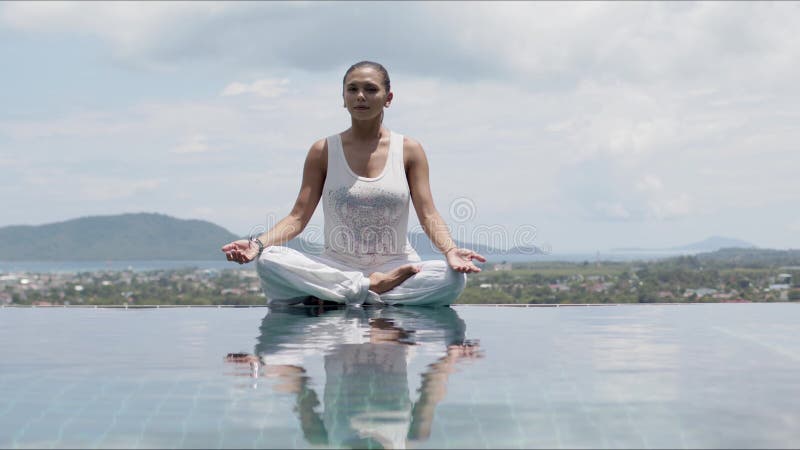 Serene woman practicing yoga in lotus posture poolside against sky