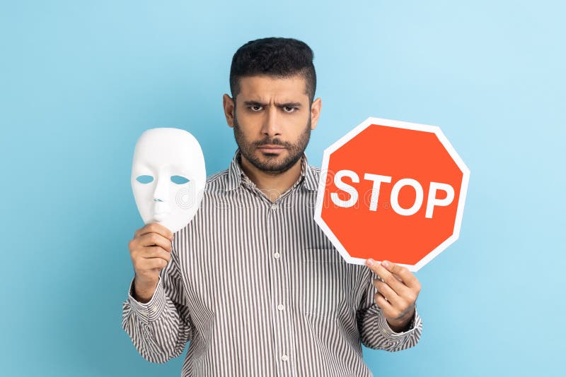 Portrait of serious businessman holding white mask with unknown face and red traffic sign, looking at camera, wearing striped shirt. Indoor studio shot isolated on blue background. Portrait of serious businessman holding white mask with unknown face and red traffic sign, looking at camera, wearing striped shirt. Indoor studio shot isolated on blue background.