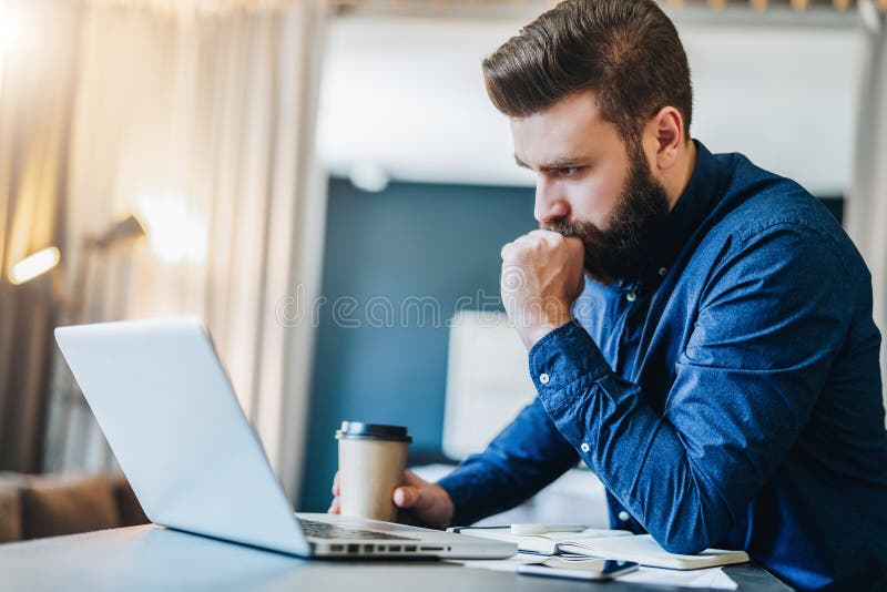 Serious bearded businessman working on computer, drinking coffee, thinking. Man analyzes information, checking email. stock photos