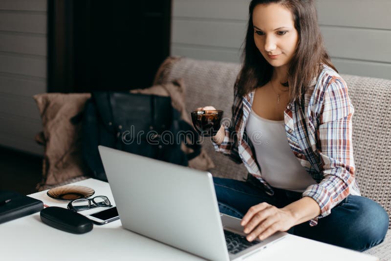 Serious beautiful brunette woman work at the notebook sit down on the sofa at home with cup on hand- check on online shops for cy