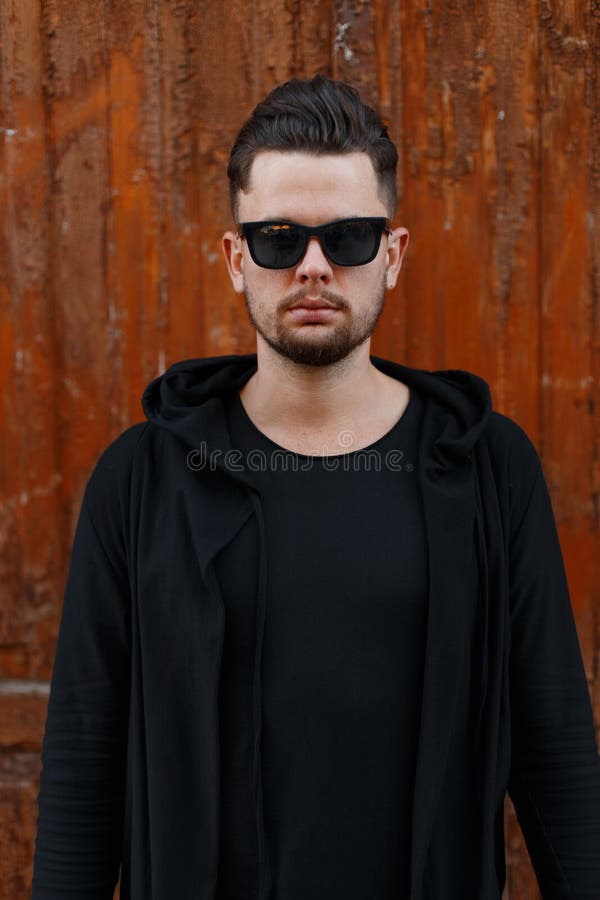 Serious brutal handsome young male hipster with fashionable hairstyle with beard in stylish sunglasses in modern black clothes royalty free stock photography Serious brutal handsome young male hipster with fashionable hairstyle with beard in stylish sunglasses in modern black clothes royalty free stock photography