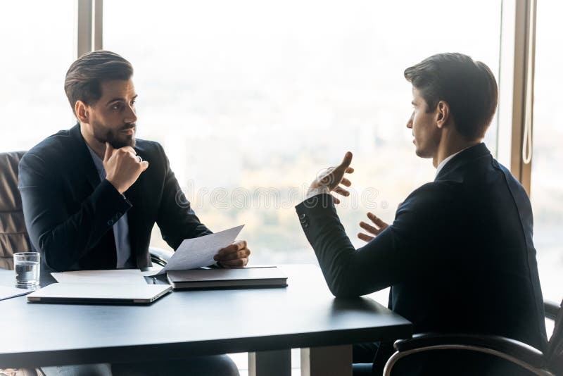 Serious businessman holding resume listening new job candidate. Young man employee holding chin thinking about decision to making deal. Boss meeting with staff stock images Serious businessman holding resume listening new job candidate. Young man employee holding chin thinking about decision to making deal. Boss meeting with staff stock images