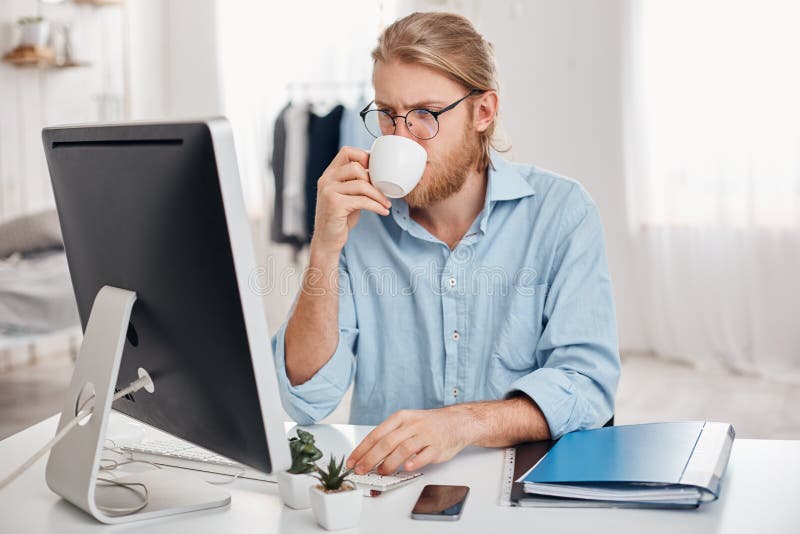 Serious concentrated on work office worker with fair hair, beard in casual outfit and glasses, prepares report, uses