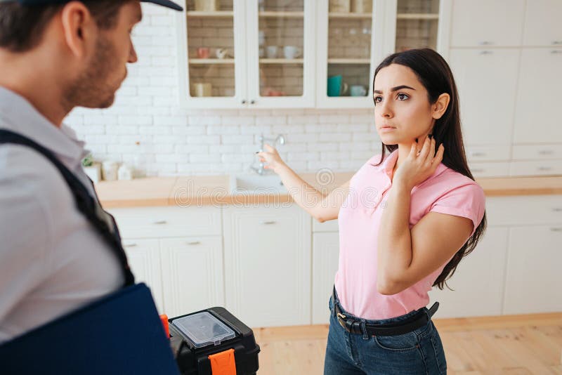 Serious concentrted woman stand with plumber in kitchen. She point on sink. Guy hold toolbox and look in that direction.