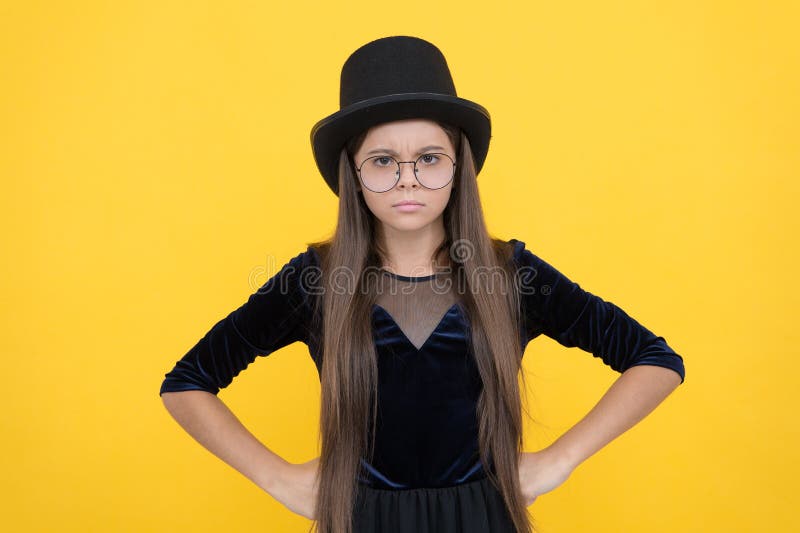 Serious female baby in glasses with long hair wear fashion party dress and hat standing with hands on hips yellow backgroud, girl.