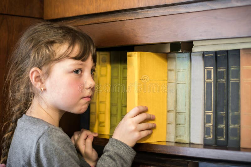 Serious Little Girl Is Taking A Yellow Book From The Bookshelf
