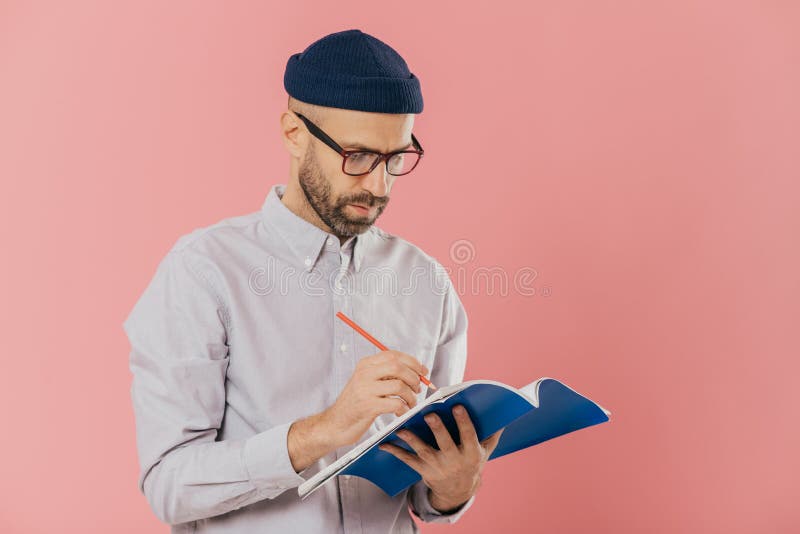 Gravi maschio scrittore scrive un saggio in blocco note, si sente ispirato, porta gli occhiali e abiti formali, preapares idee creative per il nuovo libro, modelli contro rosa parete in studio.