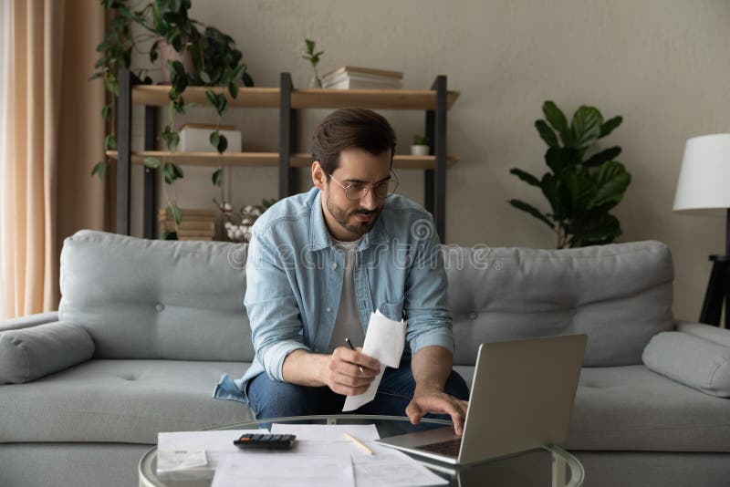 Serious millennial male in glasses focused on paying utility bills taxes rental charges online using laptop. Young man working from home office sit on couch hold paper invoice check information online. Serious millennial male in glasses focused on paying utility bills taxes rental charges online using laptop. Young man working from home office sit on couch hold paper invoice check information online