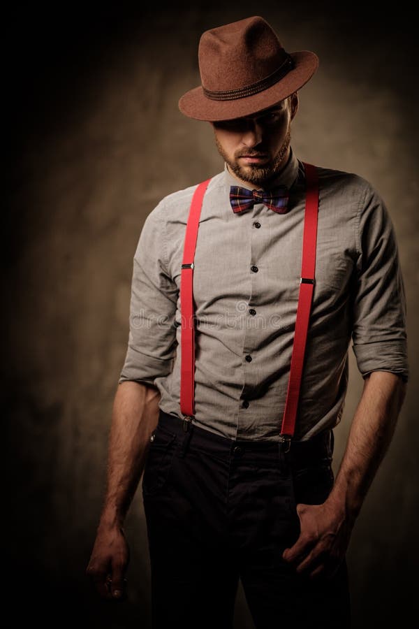 Serious old-fashioned man with hat wearing suspenders and bow tie, posing on dark background.