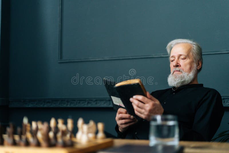 Serious senior gray-haired older male reading paper book at home sitting at wooden table with chess board, relax and