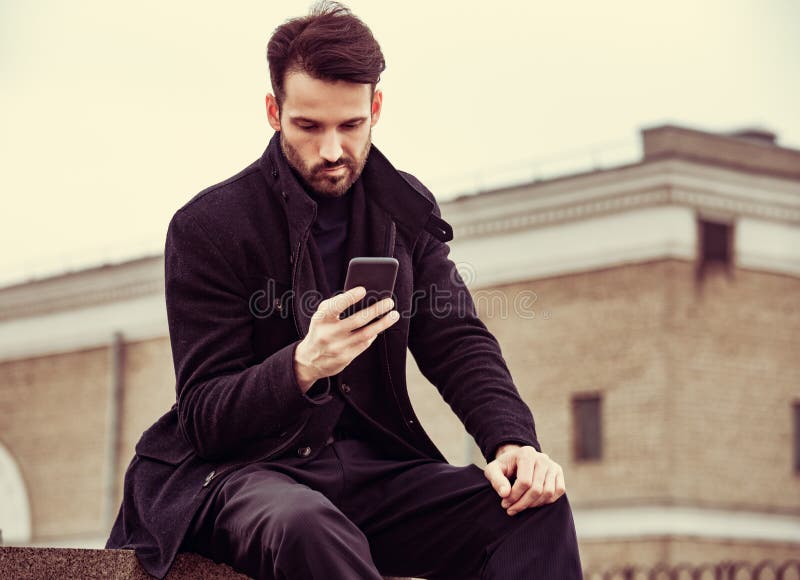 Serious thinking business man in fashion clothing texting sms looking on mobile phone in the hand outdoors autumn background. Portrait