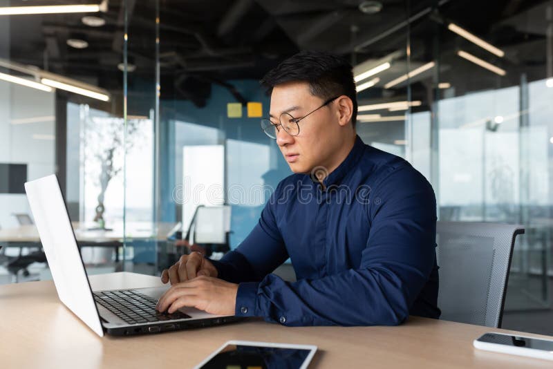 Serious and thoughtful businessman working inside office sitting at table using laptop at work, mature asian boss in