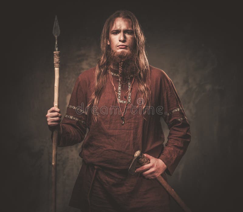 Serious viking with a spear in a traditional warrior clothes, posing on a dark background.