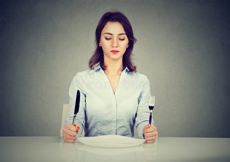 Serious woman with fork and knife sitting at table with empty plate