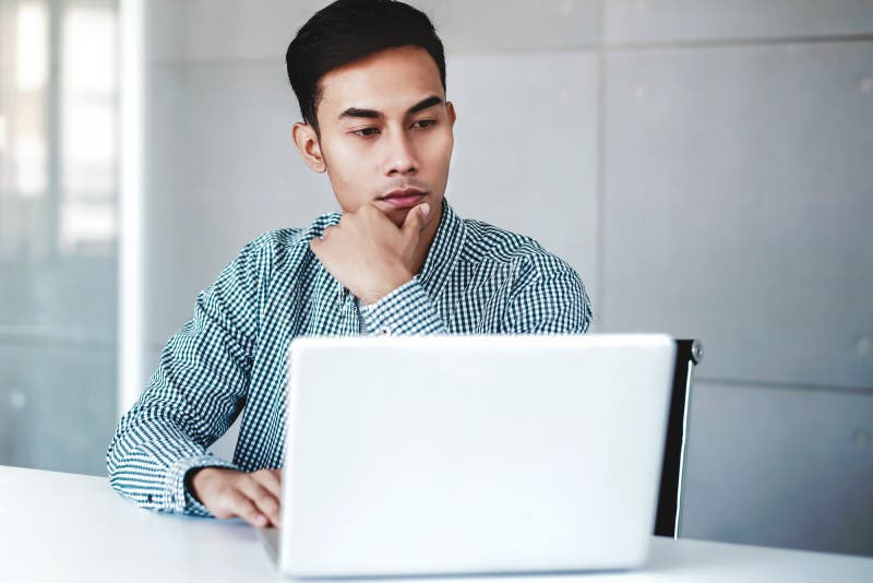 Seriously Young Businessman Working on Computer Laptop in Office. Hand on Shin, Sitting on Desk with Thoughtful Posture. royalty free stock photo Seriously Young Businessman Working on Computer Laptop in Office. Hand on Shin, Sitting on Desk with Thoughtful Posture. royalty free stock photo