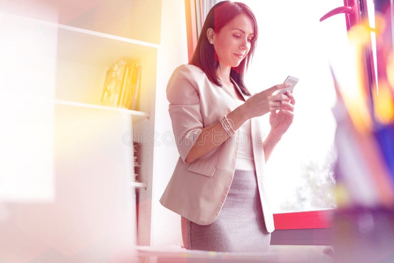 Service de mini-messages de femme d'affaires sur le smartphone par la fen?tre au bureau image stock Service de mini-messages de femme d'affaires sur le smartphone par la fen?tre au bureau image stock