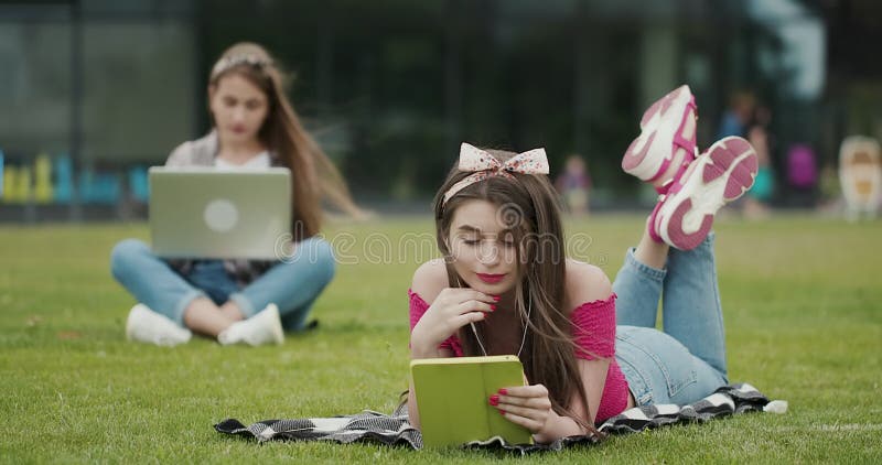 Sesión dos femenina en el césped de un parque precioso y utilizar un ordenador del dispositivo para el ordenador portátil y la tab