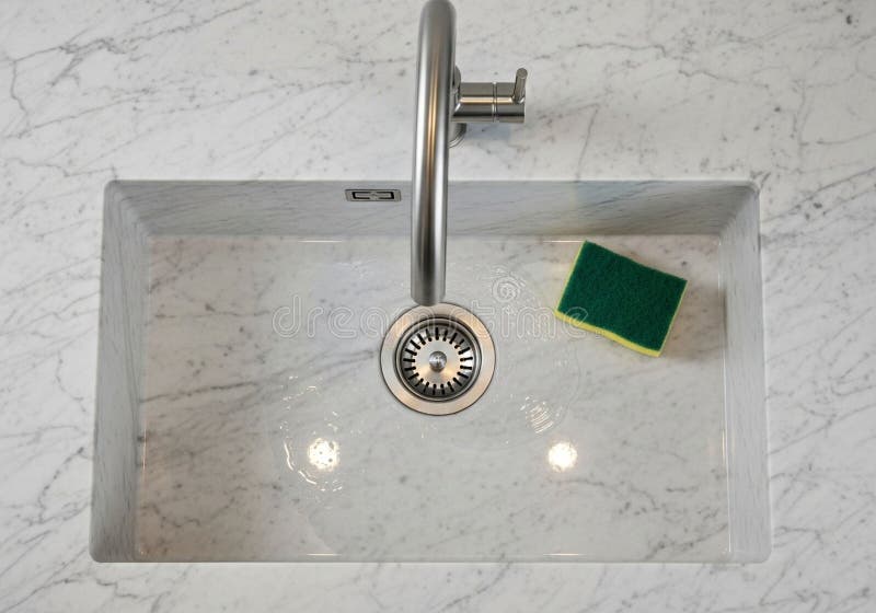 A rectangular stainless steel kitchen sink set into a white marble countertop with gray veining. The sink features a modern, curved faucet mounted centrally, and a drain with a strainer. A two-tone sponge, green on the top and yellow on the bottom, is placed on the right side of the sink. The marble surface adds elegance and luxury to the overall design. The setup is clean and minimalist, ideal for a contemporary kitchen setting. Sink ideal illustrations