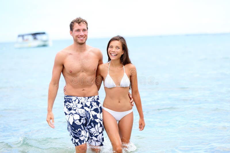 Beach couple in love walking happy in water holding hands around eachother enjoying summer holidays vacation travel. Multiracial young couple, Asian woman, Caucasian men in their 20s. Beach couple in love walking happy in water holding hands around eachother enjoying summer holidays vacation travel. Multiracial young couple, Asian woman, Caucasian men in their 20s.