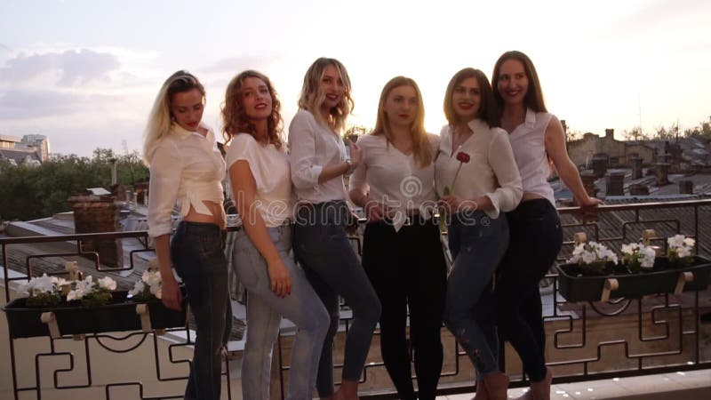 Sex caucasian flickor står på en balkong och poserar för kameran Tillfällig kläder Slutet upp av flicka` s lägger benen på ryggen