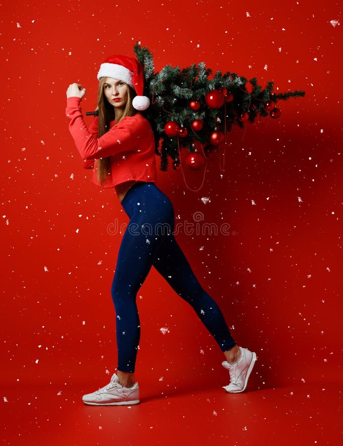 Sexig kvinna för julkonditionsport som bär den santa hatten som rymmer xmas-trädet på hennes skuldror snowflakes arkivfoto Sexig kvinna för julkonditionsport som bär den santa hatten som rymmer xmas-trädet på hennes skuldror snowflakes arkivfoto