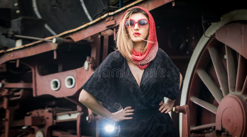 attractive girl with red head scarf and sun glasses posing on the platform in front of a vintage train. Woman vintage