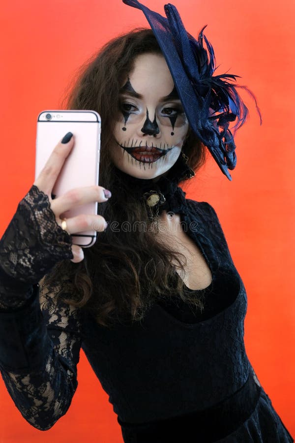 Halloween makeup for a party. girl in a black dress and hat takes pictures of herself on the phone. A woman makes a royalty free stock photo Halloween makeup for a party. girl in a black dress and hat takes pictures of herself on the phone. A woman makes a royalty free stock photo