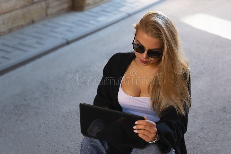 Sexy blonde in sunglasses works at the computer while sitting on the pavement hiding in the shade from the scorching sun