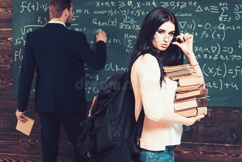 Sexy brunette female student holding her glasses while carrying pile of books. Young college girl in pink cardigan walking in front of green board.