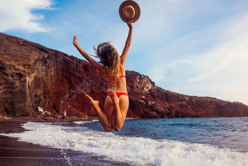 Sexy Frau in den Bikiniwegen auf rotem Strand im santorini auf dem Seeweg. das Mädchen springt, die Arme Auslösen von, die Spaß So Sexy Frau in den Bikiniwegen auf rotem Strand im santorini auf dem Seeweg. das Mädchen springt, die Arme Auslösen von, die Spaß So