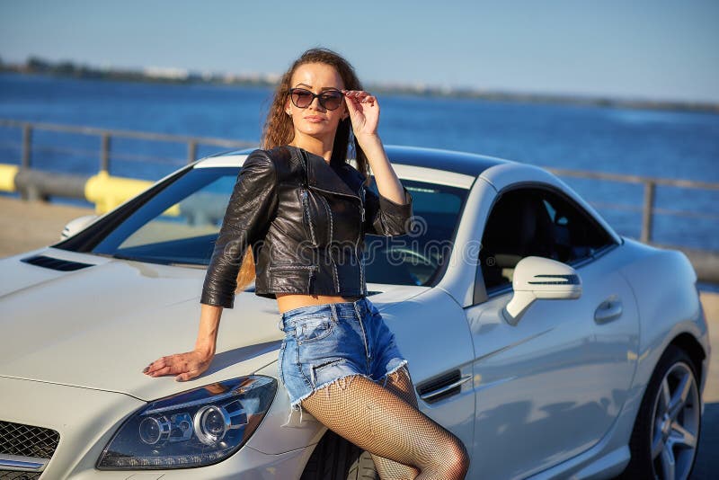 sexy girl in a leather jacket, denim shorts and black pantyhose in a grid stands near the car.