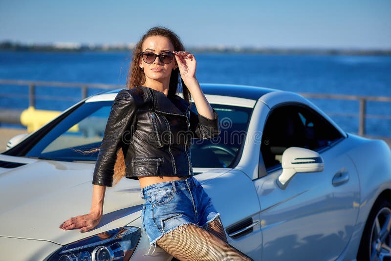 sexy girl in a leather jacket, denim shorts and black pantyhose in a grid stands near the car.