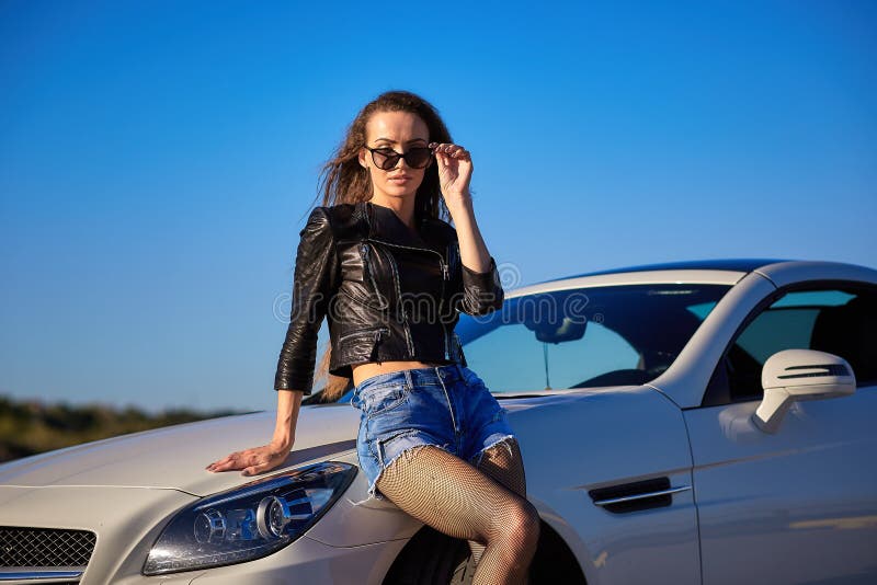 sexy girl in a leather jacket, denim shorts and black pantyhose in a grid stands near the car.