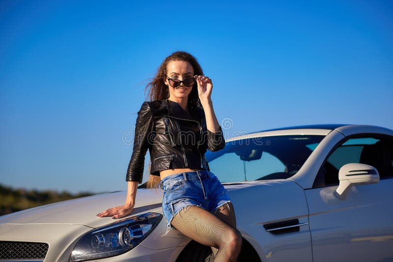 sexy girl in a leather jacket, denim shorts and black pantyhose in a grid stands near the car.