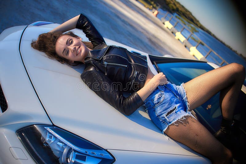 sexy girl in a leather jacket, denim shorts and black pantyhose in the net lies on the hood of the car.