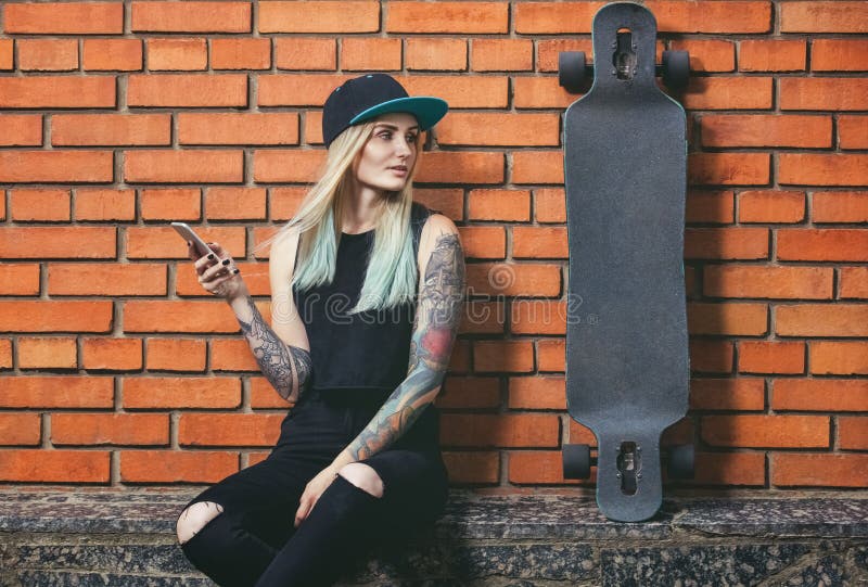 hipster girl in tattoo against a red brick wall with a long board.