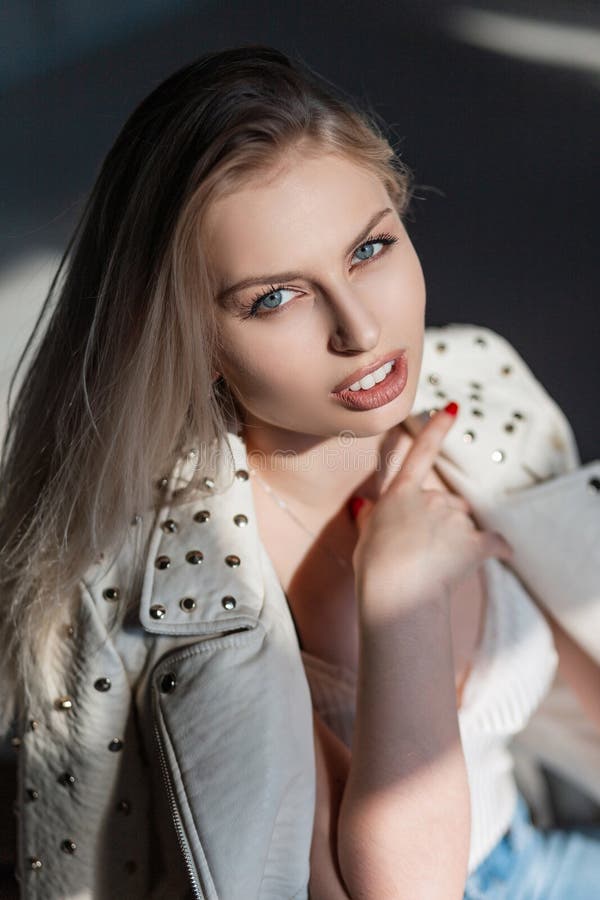 Sexy pretty attractive young blond woman with natural make-up in a stylish jacket with metal rivets in a room on a sunny day.