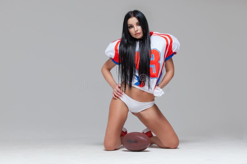 Sexy sporty brunette dressed in bikini and American football uniforms and jersey T-shirt sitting on the floor with a ball grey background. Sexy sporty brunette dressed in bikini and American football uniforms and jersey T-shirt sitting on the floor with a ball grey background