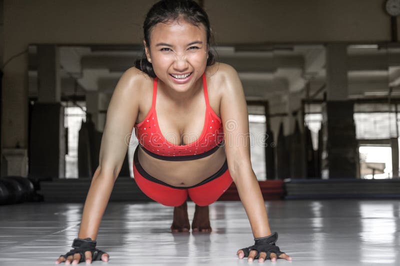 sweaty Asian woman in sport clothes doing push ups on gym dojo floor smilin...