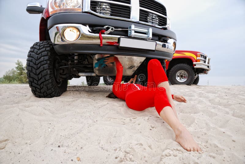 A driver is lying under the off-road vehicle in the dunes. She is repairing the car. Young woman is wearing a red catsuit. She is barefooted. A driver is lying under the off-road vehicle in the dunes. She is repairing the car. Young woman is wearing a red catsuit. She is barefooted.
