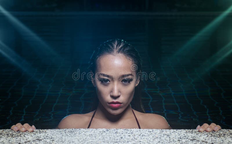 Closeup face of sensual Asian woman with wet hair standing in the swimming pool and looking into the camera during summer evening over blue and green lights reflections background. Closeup face of sensual Asian woman with wet hair standing in the swimming pool and looking into the camera during summer evening over blue and green lights reflections background